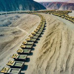 20 lines and 100 number of  mining dump trucks  located middle of the picture  and left and right side is a mountains. the huge truck in middle of them has green lights and cargo and body that resembles like  the robots in the Transformers movie