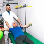 یک بیمار که برای معالجه در یک کلینیک فیزیوتراپی مشغول انجام تمرینات فیزیوتراپی است و فیزیوتراپیست در حال کمک به وی است. این بیمار در حال راه رفتن به کمک فیزیوتراپیست است. این بیمار یک مرد مسن است که در حال رضایت از انجام این کار است.