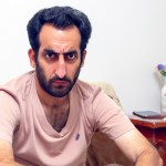 تصویر یک مرد جوان ایرانی با زیر پیراهنی کهنه که در خانه نشسته و ناراحت است