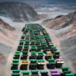 100 number of mining dump trucks located middle of the picture and left and right side is a mountains. the huge truck in middle of them has green lights and cargo and body that resembles like the robots in the Transformers movie