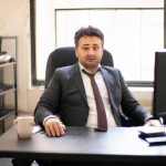 یک کارمند چهل ساله، پشت میز قهوه‌ای تیره کا روی آن مانیتور مشکی هست، پشت سرش یک پنجره بزرگ رو به دیوار سفیدرنگ. لباس سوره ای و قهوه‌ای چهارخانه متوسط. چشم و مو مشکی، فضای ایرانی.