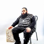 بهنام هاشمی یک مردی با قد 165 سانتیمتر چاق با لباس مشکی دارای ریش سیاه و سفید نشسته روی چهارپایه پلاستیکی