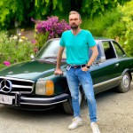 یک مرد 40 ساله با تیشرت سبز و ریش و شلوار جین آبی در کنار یک ماشین بنز قدیمی  در یک باغ پر گل