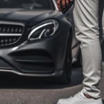 یک هسمتر که یک گوشی آیفون ۱۵ پرو مکس دستش هست و در حال راه رفتن با بنز ای ام جی