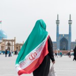 The girl wearing hijab and Islamic clothing, holding the flag of Iran, is moving towards Imam Reza's shrine from behind.