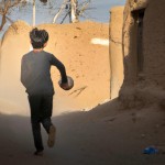 علی بیرانوند دروازه بان پرسپولیس در حال دویدن با توپ پارچه‌ای در میان کوچه‌های خاکی روستای سربند. اطرافش خانه‌های ساده روستایی با دیوارهای گلی و چند درخت پراکنده دیده می‌شوند. علی پسری ۷-۸ ساله با موهای مشکی کوتاه و لباس‌های ساده محلی است