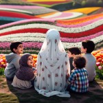 مادر محجبه پشت به دوربین نشسته.چادر سفید .۲تا پسر ۴ ساله‌ و ۵ساله و ۲ تا دختر ۲ساله و ۷ساله‌ش دورِ مامان حلقه زدن و به مامان نگاه می‌کنن. زیرشون چمن و روبه روشون منظره‌ای از یک دشت لاله