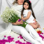 یک تصویر بساز از یک مادر 50 ساله ایرانی با چادر سفید که در یک مکان نورانی نشسته است و کودک دخترش را در آغول گرفته است و زیر پاهایش پر از گل های رنگی است