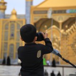 پسر عکاس مذهبی حین عکاسی از حرم امام رضا. لباس مشکی و جفیه طلایی رنگ