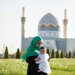 یک عکس مادر با چادر سفید و فرزند یک ساله در بغل  اطراف آن چمن و پشت آن گنبد امام رضا باشد