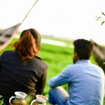 عکس یک مرد ویک خانوم که پشتشان به ماست ودر مزرعه ای  سرسبز کنار یک قایق چوبی به گل نشسته در حال چایی خوردن  با صورت های تار تولید کن
