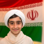 عکس یک پسر ۱۲ ساله ایرانی که لباس روحانیت ایرانی پوشیده یعنی عبای قهوه ای ساده پوشیده و عمامه سفید بر سر دارد و با لبخند به دوربین نگاه میکند و در پس زمینهتصویر مات پرچم ایران هست تولید کن