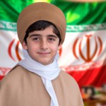عکس یک پسر ۱۲ ساله ایرانی که لباس روحانیت ایرانی پوشیده یعنی عبای قهوه ای ساده پوشیده و عمامه سفید بر سر دارد و با لبخند به دوربین نگاه میکند و در پس زمینهتصویر مات پرچم ایران هست تولید کن