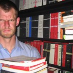 Convert this image into a black-and-white photograph of the handsome man shown in the reference photo (use the reference face for the uploaded image). His head is tilted back in a moment of deep creative thought or awe. He wears a loose, open-collared white shirt, standing in a dimly lit study filled with stacks of books and papers. The image is captured with a slow shutter speed, creating dramatic horizontal motion blur that softens the background and the edges of his form
