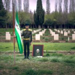 عکس شهید قاسم سلیمانی با لباس نظامی که در دستش پرچم ایران را گرفته و در گلزاری ایستاده به صورت واقعی در روز باشد
