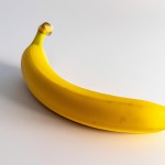 “A fresh, bright yellow banana with smooth and shiny peel placed on a plain white background. Soft light shines from the left side, casting a gentle shadow beneath the banana. The banana is slightly curved and looks natural, with visible texture on its peel.”