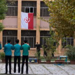 حیاط مدرسه ایرانی با پرچم جمهوری اسلامی و دانش آموزان پسر در حال خواندن سرود همگانی