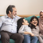 A joyful Iranian family gathered inside a simple and cozy living room, horizontal composition, middle-aged father and mother sitting comfortably on a modest sofa, laughing and talking warmly with their children, natural candid interaction, genuine smiles and eye contact, children gathered around them including a small child, a teenage boy, a teenage girl, and young adult son and daughter, females wearing full hijab and modest clothing, culturally appropriate appearance, simple home decor, soft